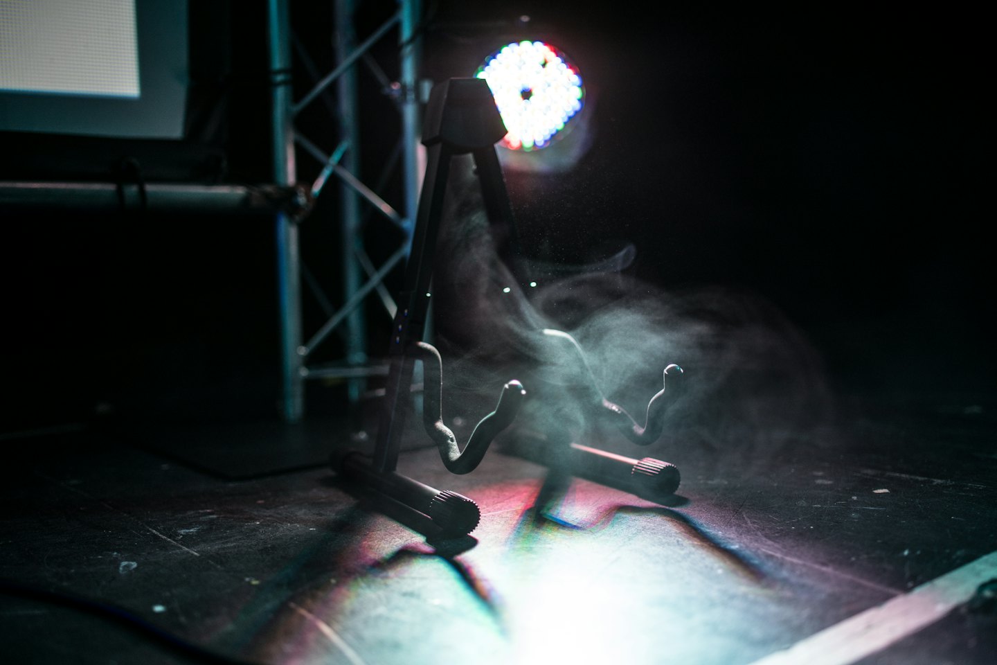 A metal guitar stand sits on a smoky stage floor.
